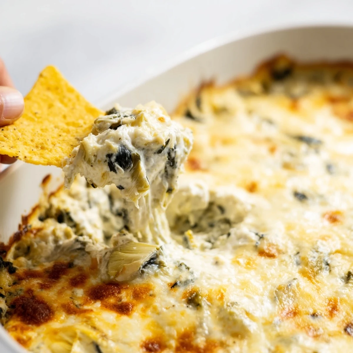 A spoon lifts a cheesy scoop of Spinach Artichoke Dip from the baking dish, revealing tender greens and melted cheese inside.
