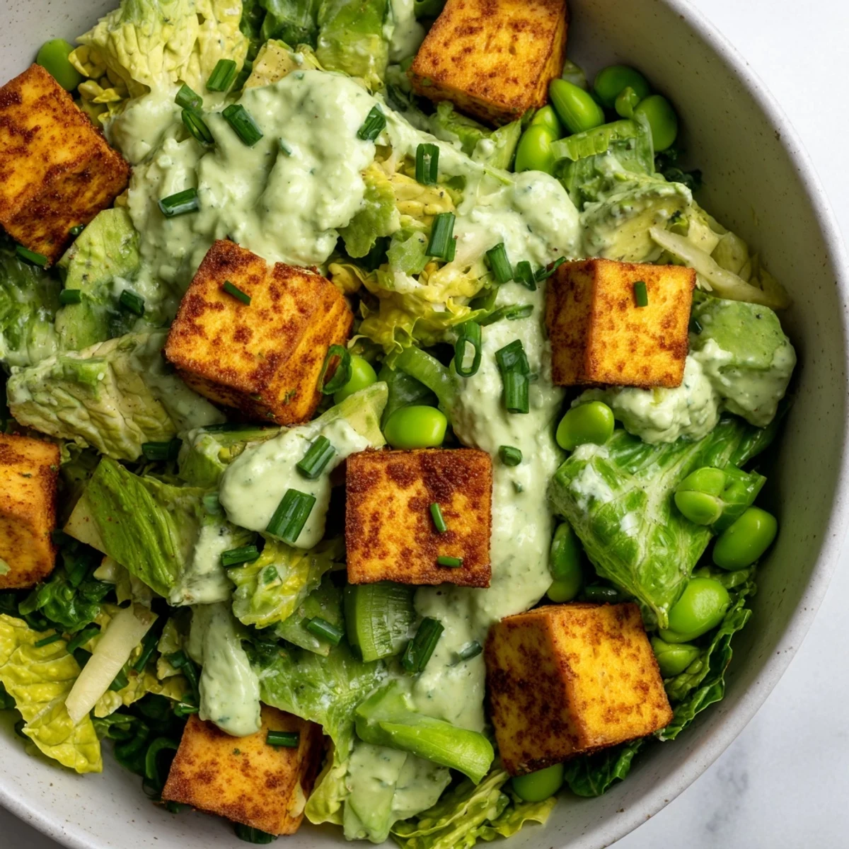 Freshly baked crispy tofu cubes sit atop a vibrant Green Goddess Salad, featuring romaine, cabbage, and creamy avocado with a green herb dressing.