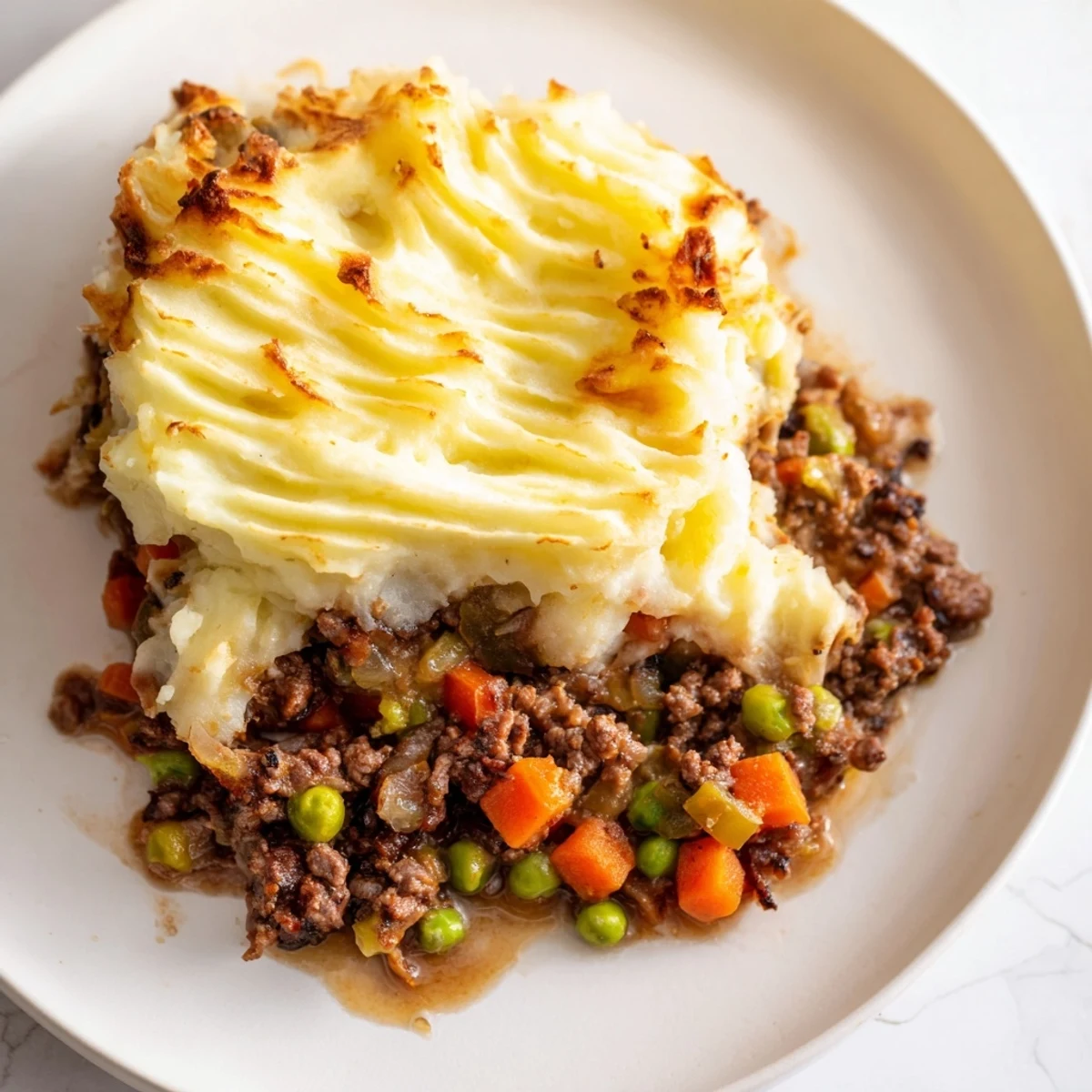 Serving of Classic Beef Cottage Pie on a plate with gravy pooling beside fluffy mash and tender beef and peas.