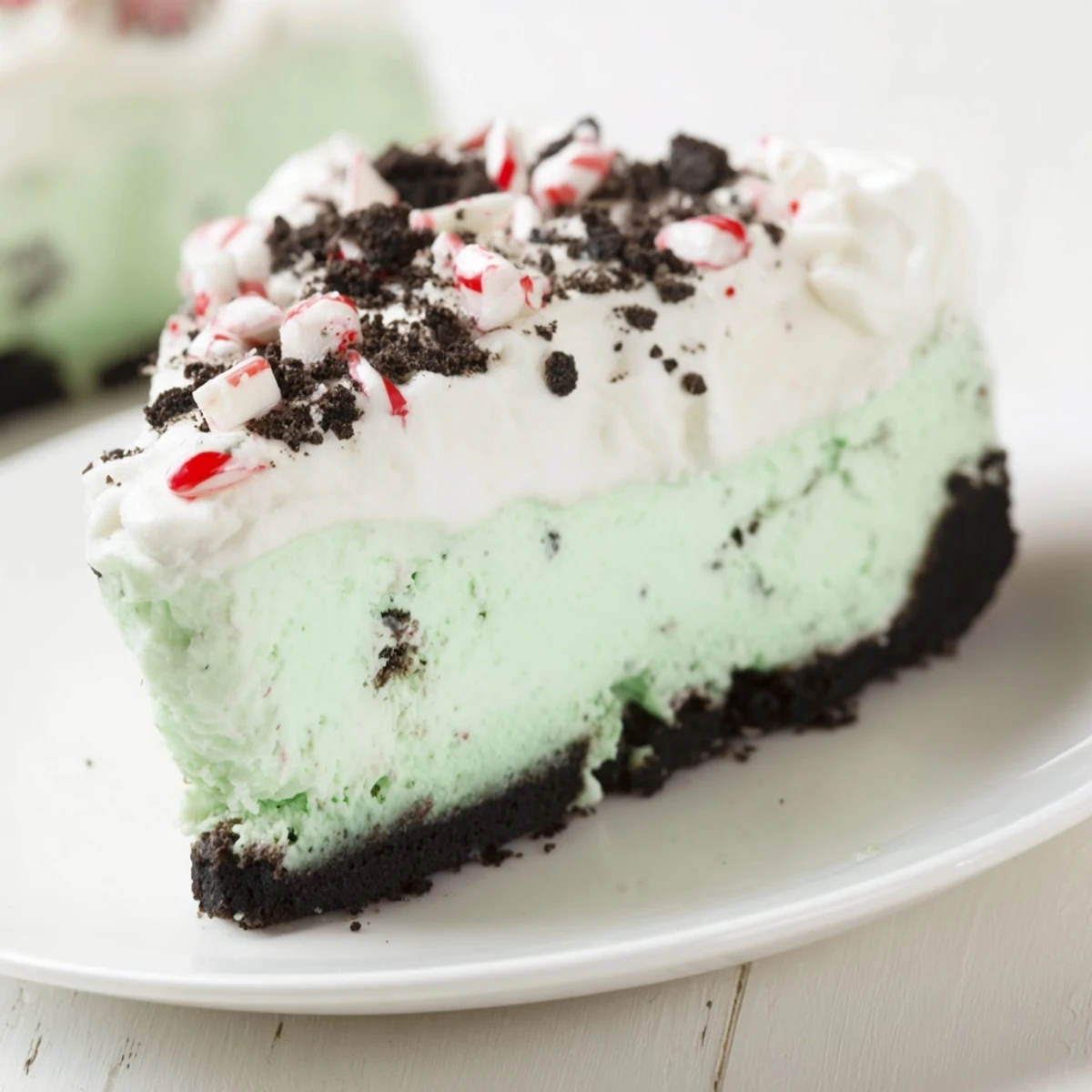 A chilled slice of Peppermint Cheesecake with Oreo Crust sits on a white plate, revealing a creamy, mint-green interior and chocolate cookie base, topped with crushed candy canes and a dollop of whipped cream.