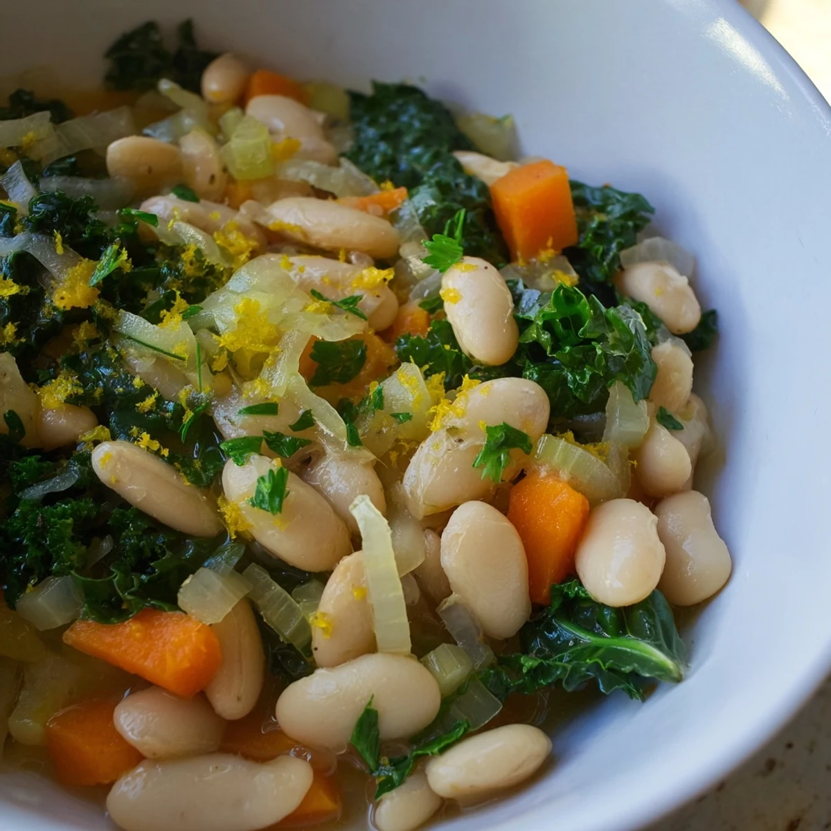 A close-up shot highlights the texture of this White Bean and Kale Soup with Lemon Zest, featuring chunky diced carrots and celery mixed with creamy white beans and ribbons of wilted kale. Steam gently rises from the surface, suggesting it is served piping hot, perhaps alongside a slice of rustic bread for dipping.