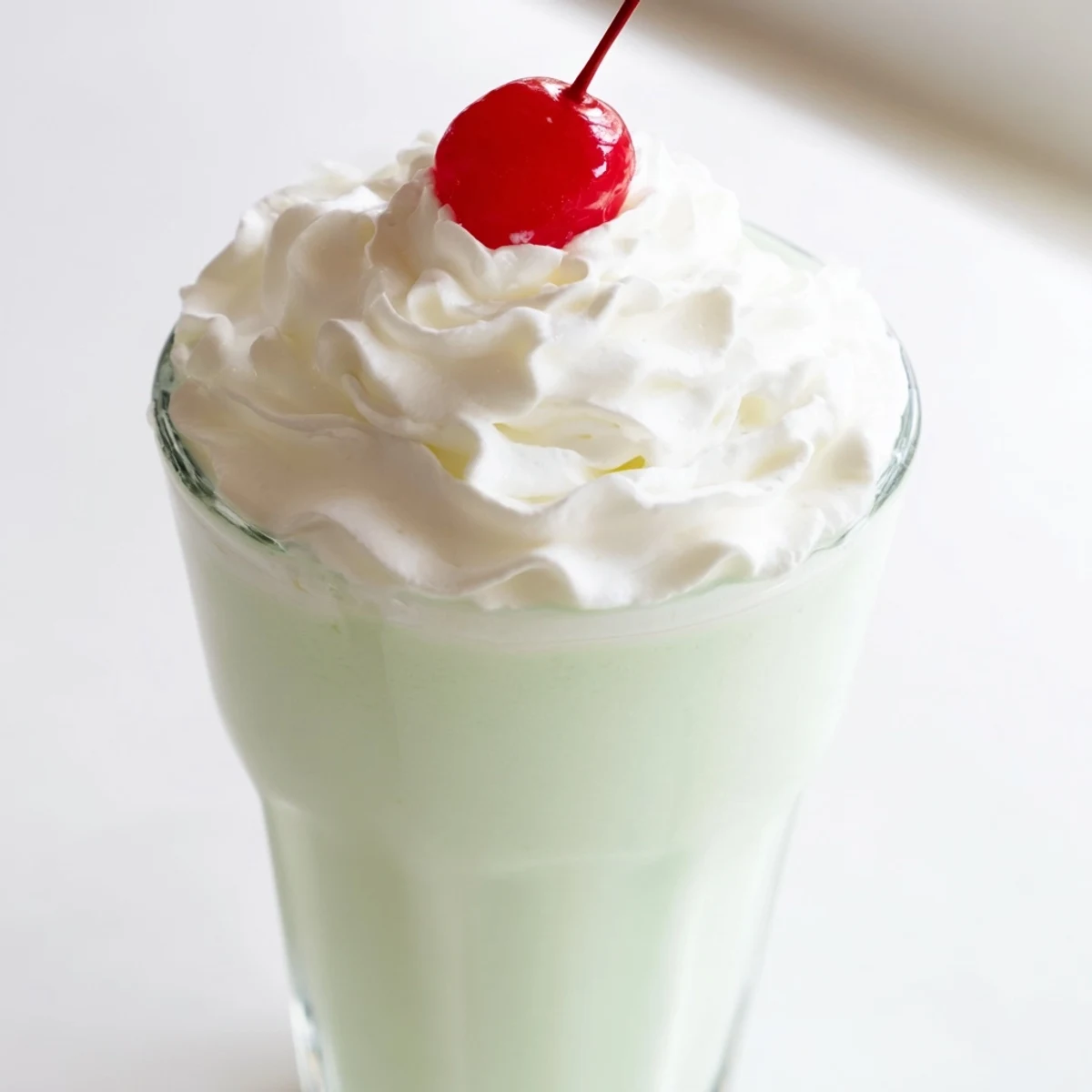 Creamy mint milkshake topped with a maraschino cherry, served in a frosted glass for a refreshing dessert.