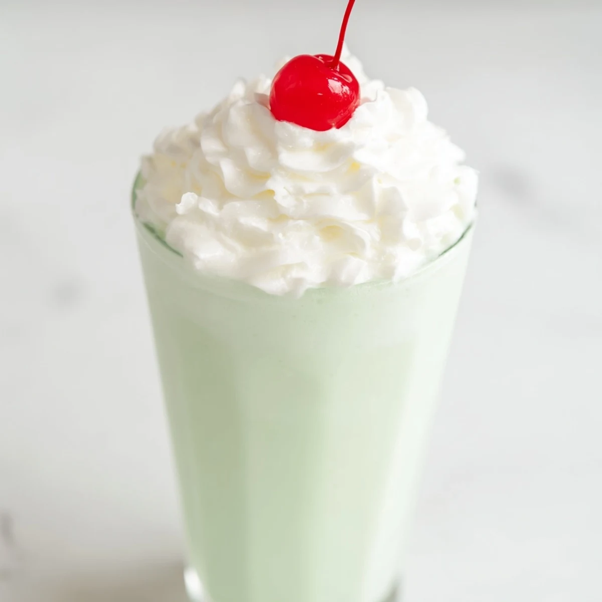 A tall mint milkshake with whipped cream and a bright maraschino cherry on a sunny patio table.