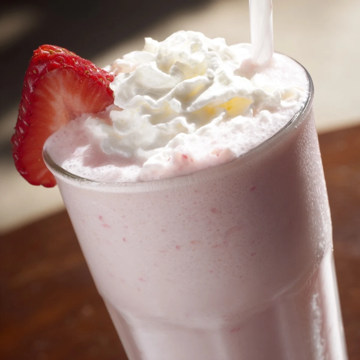 Glass of delicious strawberry milkshake topped with whipped cream and fresh strawberries on a wooden table.  