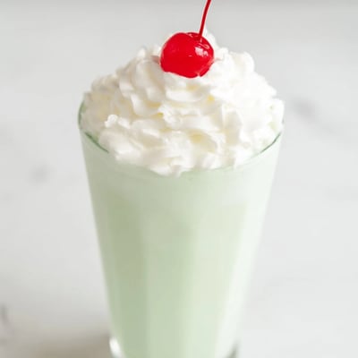 A tall mint milkshake with whipped cream and a bright maraschino cherry on a sunny patio table.