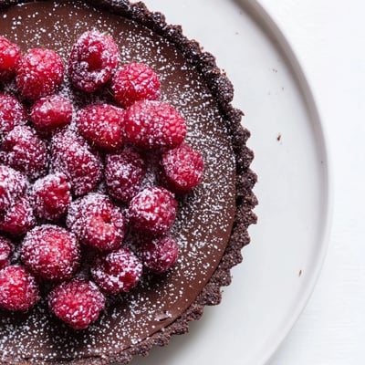 A sliced chocolate raspberry tart reveals glossy ganache and tart raspberries, dusted with powdered sugar, ready for a festive dessert table.